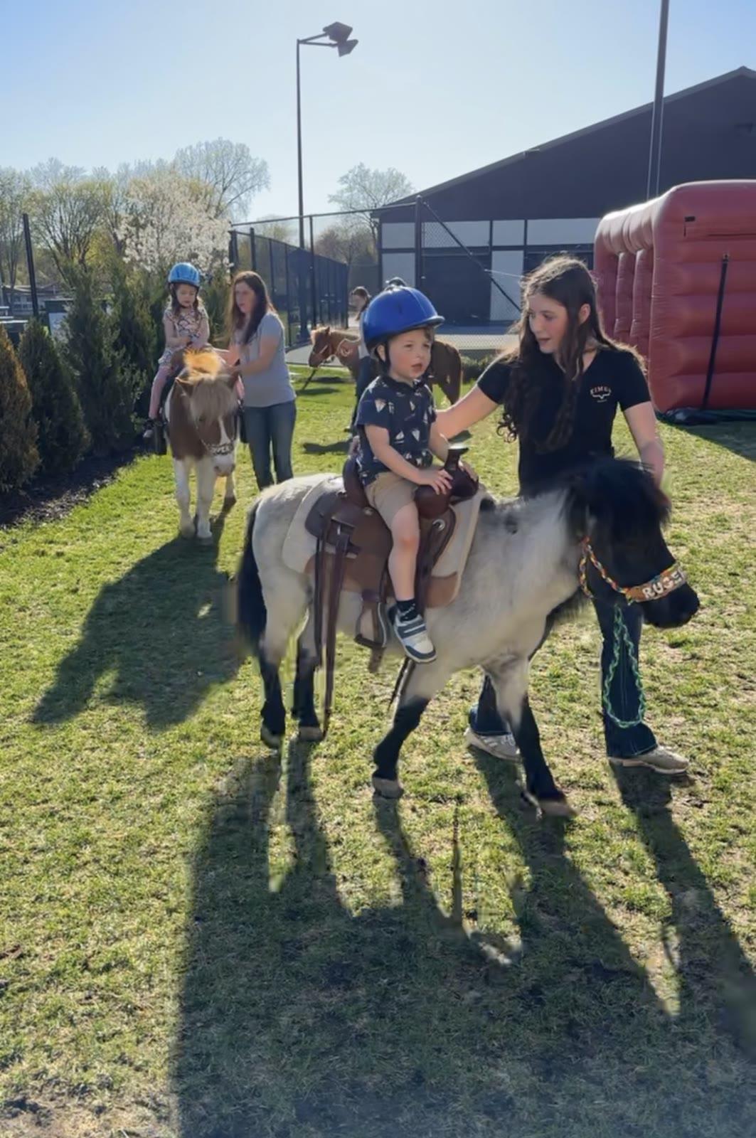 A child in a helmet sits on a pony held by a young woman on a lawn near a large building and inflatable structure.