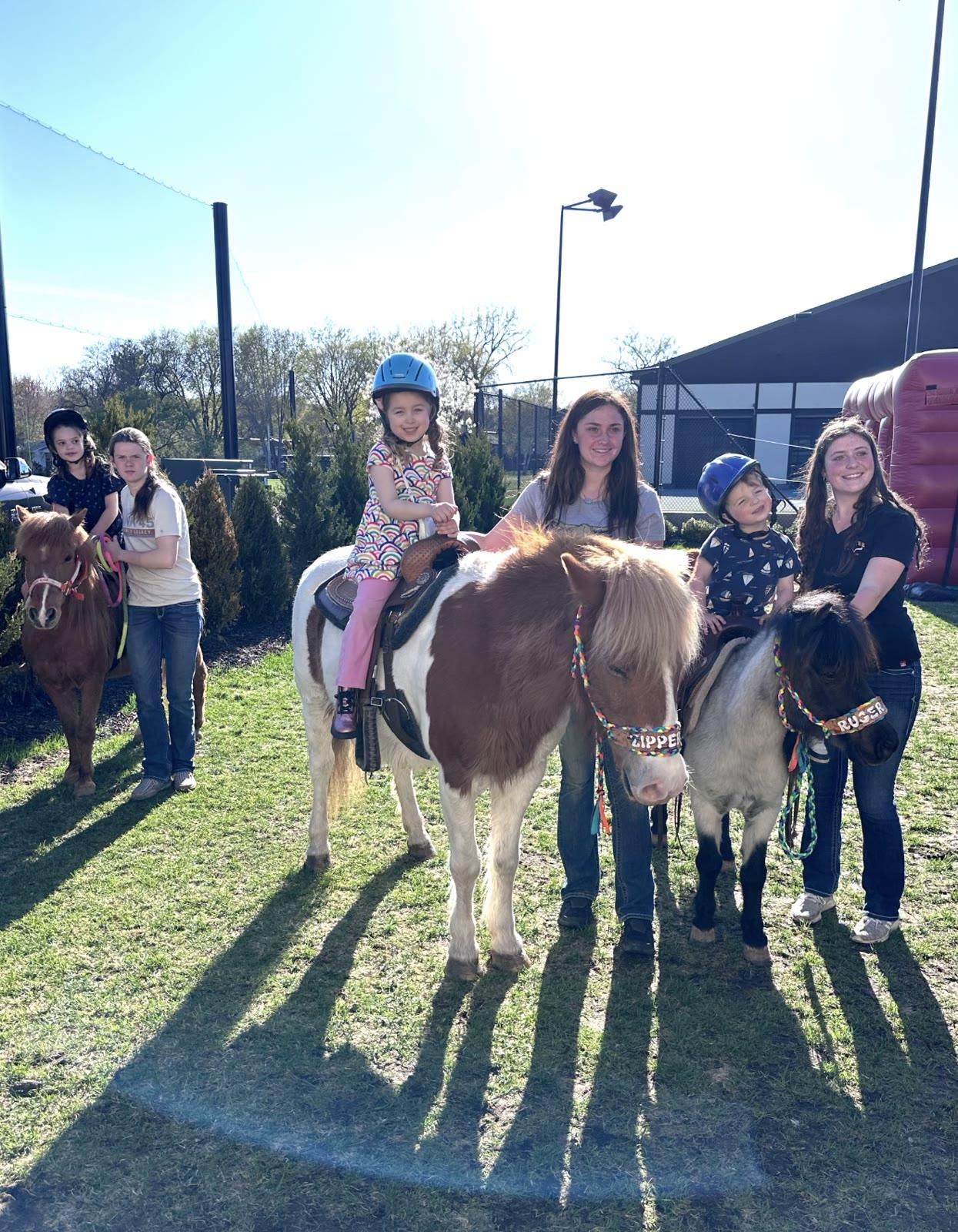 Two children ride ponies named Zipper and Ruger while two handlers stand beside them near a large building.