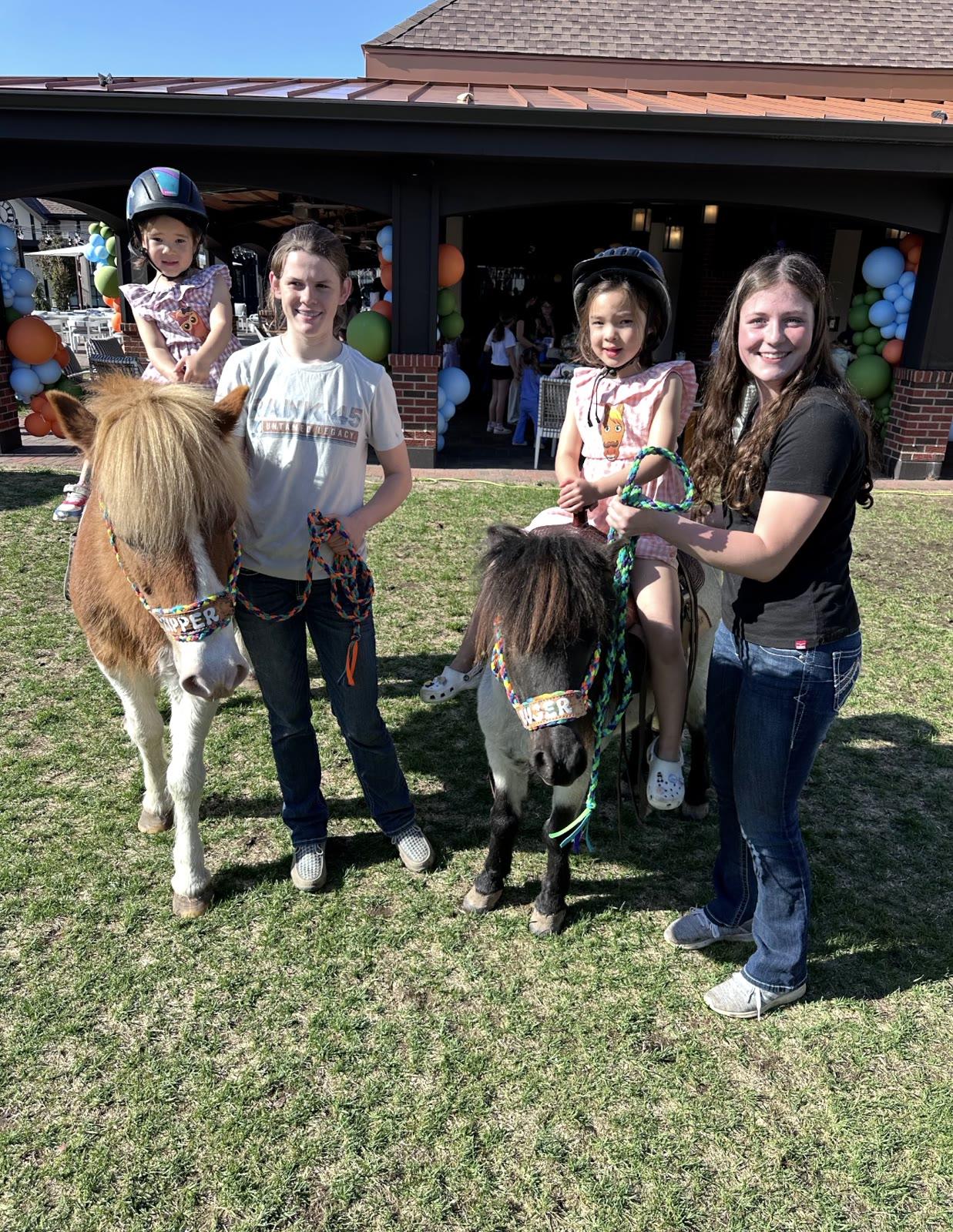 Two women hold the ponies Zipper and Ruger with children nearby under a patio decorated with balloon garlands.
