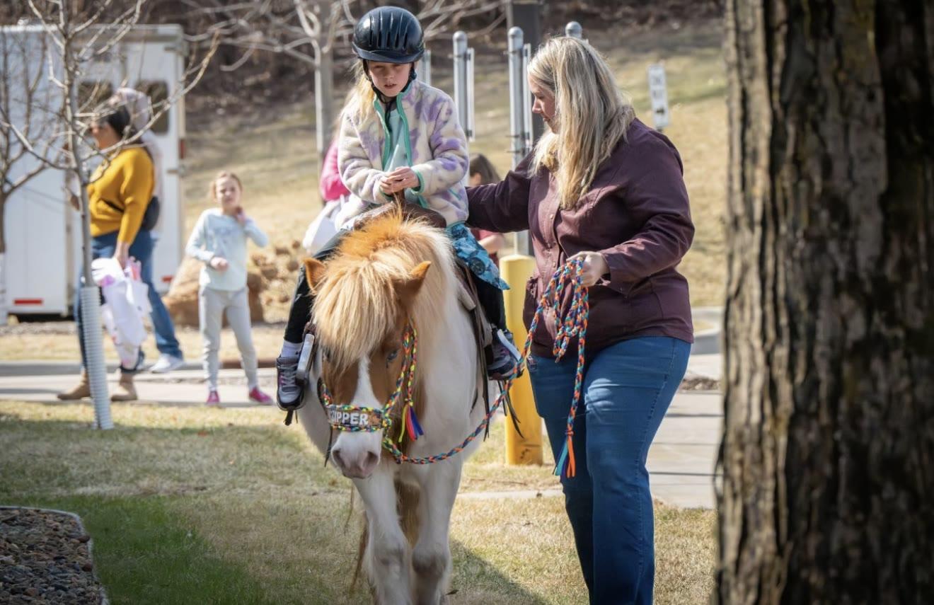 A woman leads the pony Zipper with a child rider along a path near a trailer and trees.