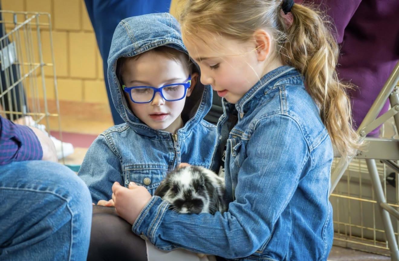 Two children in matching denim jackets sit together, one holding a black-and-white rabbit, inside a room with a wire pet crate and cinderblock wall.