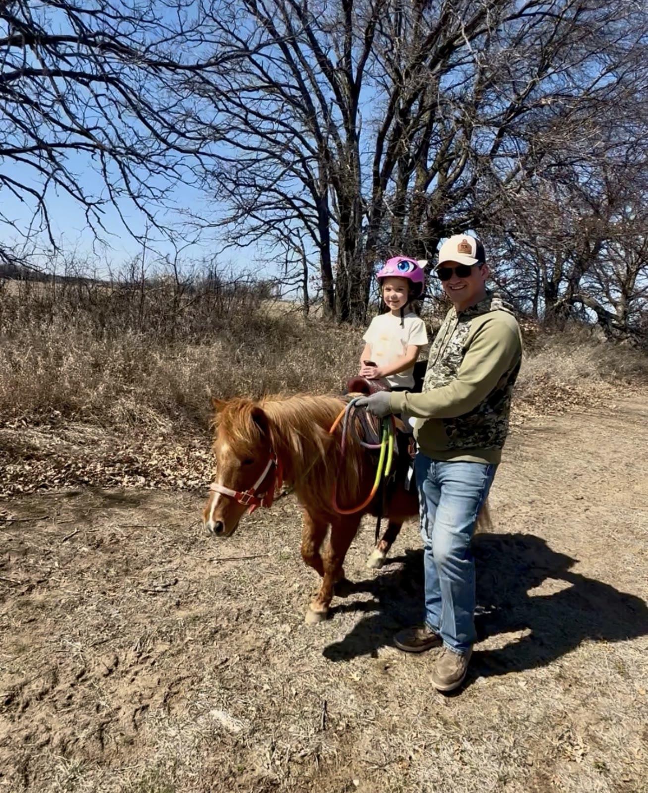 A man in a camo hoodie leads a small chestnut pony carrying a young girl in a pink helmet along a dirt path lined with bare trees and dry brush.