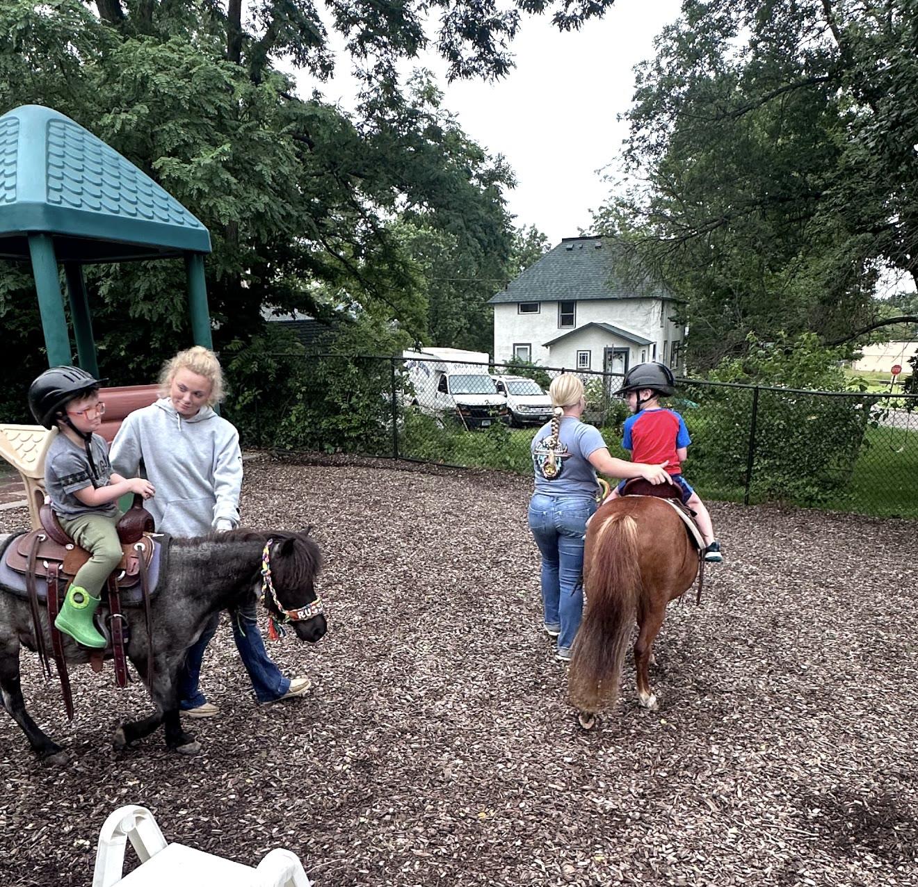 Two young riders in helmets on ponies led by handlers across a playground.