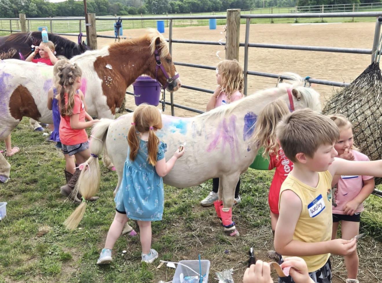 Two staff members hold the lead ropes of two ponies while a group of young children gather around them at an outdoor playground with a gazebo.