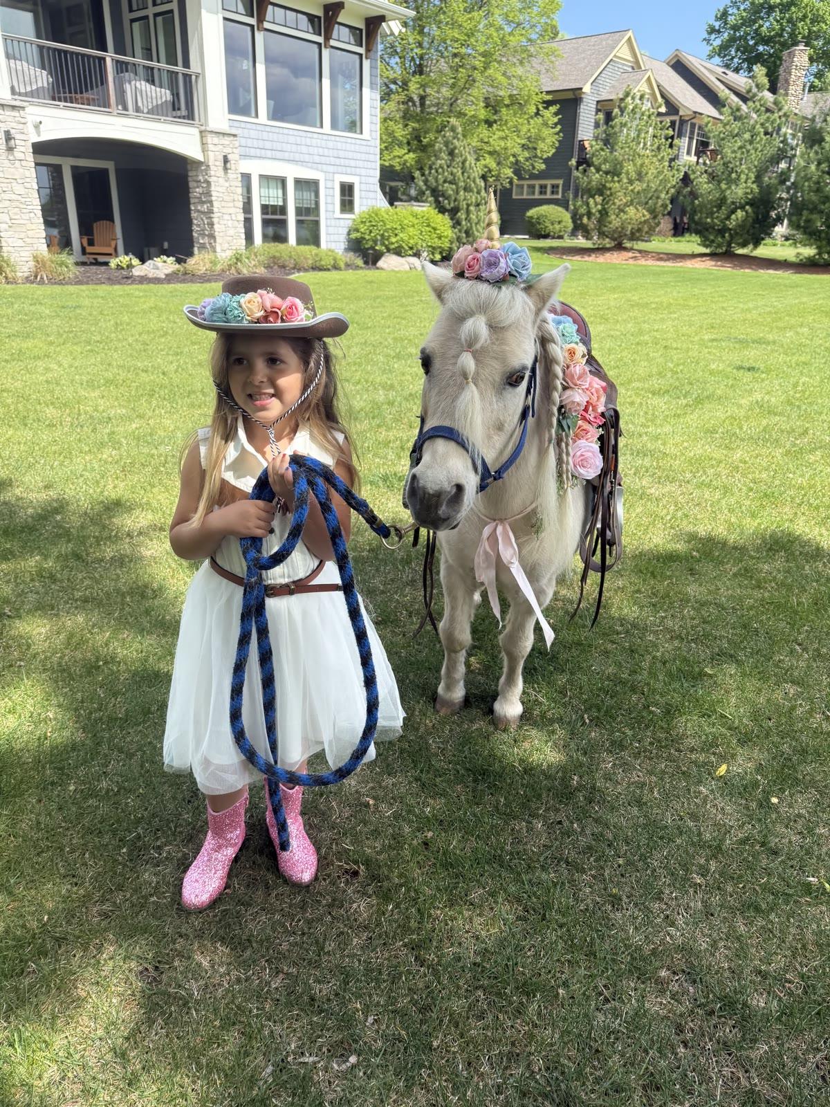 A young girl in a white dress and flower-trimmed cowboy hat holds the lead rope of a pony decorated with a flower crown and braided mane, on a lawn in front of a large house.