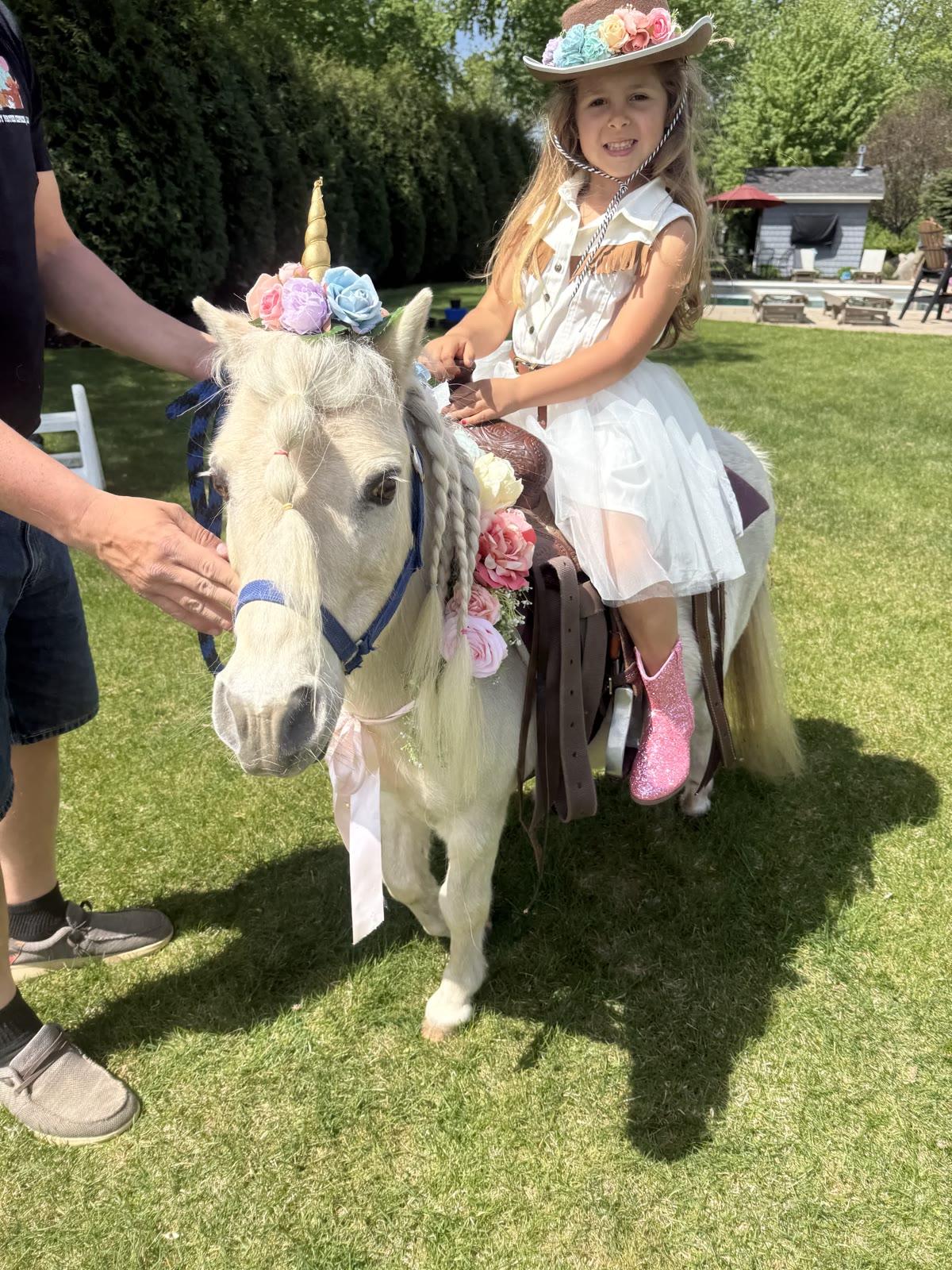 A man helps a girl in a cowboy hat sit on a pony decorated with a flower crown, on a lawn near a hedge, with white folding chairs nearby.