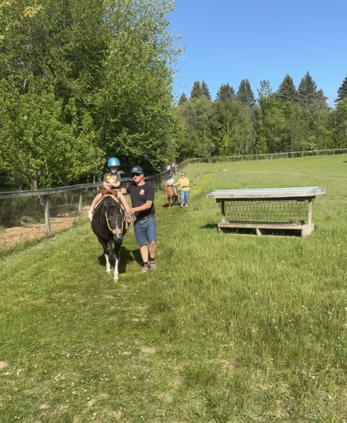 A man leads a black-and-white pony carrying a boy in a blue helmet along a grass path in a fenced pasture, with a wooden hay feeder and other riders behind them.