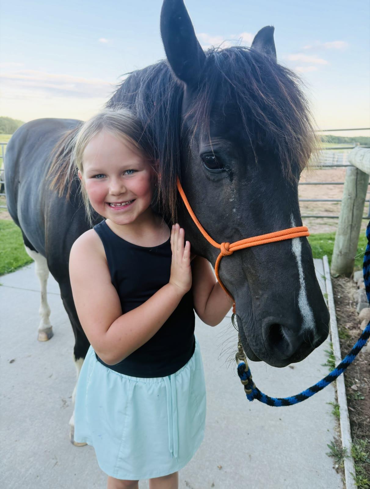 A young girl smiles while resting her hand on the face of a dark pony named Nellie, standing on a paved path near a pasture fence.