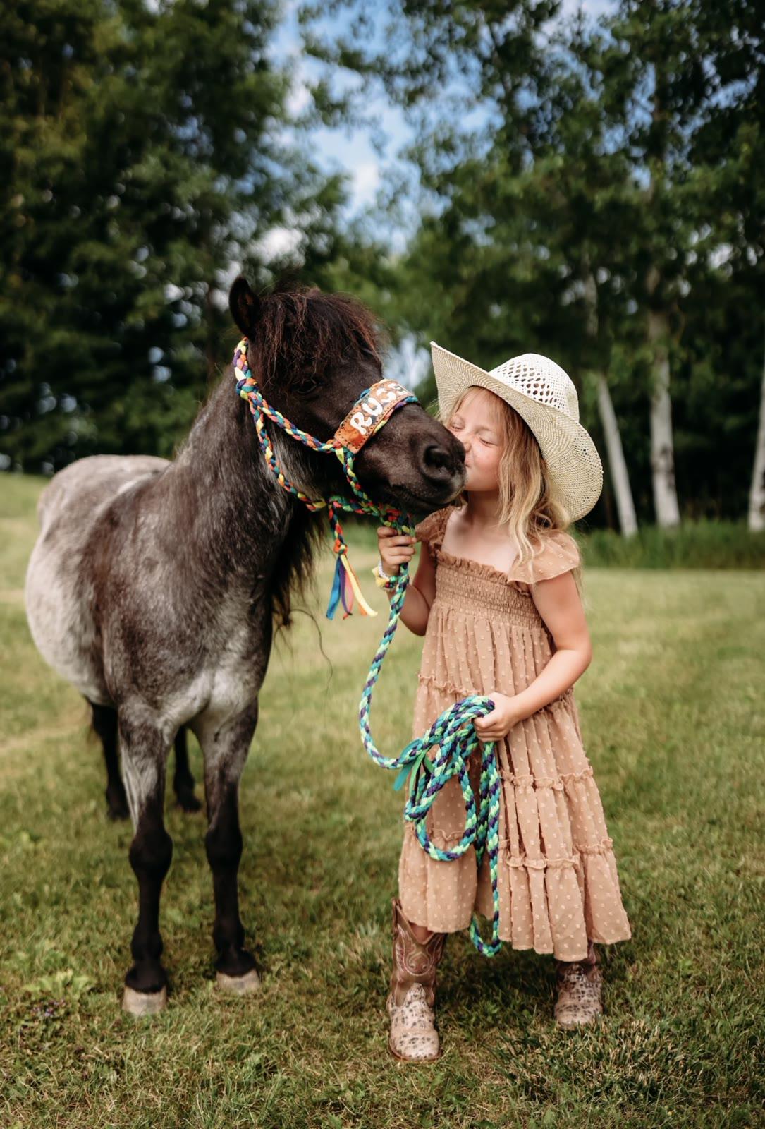A girl in a straw hat and boots leans in to touch noses with a gray pony named Ruger, wearing a colorful braided halter, in a grassy field.