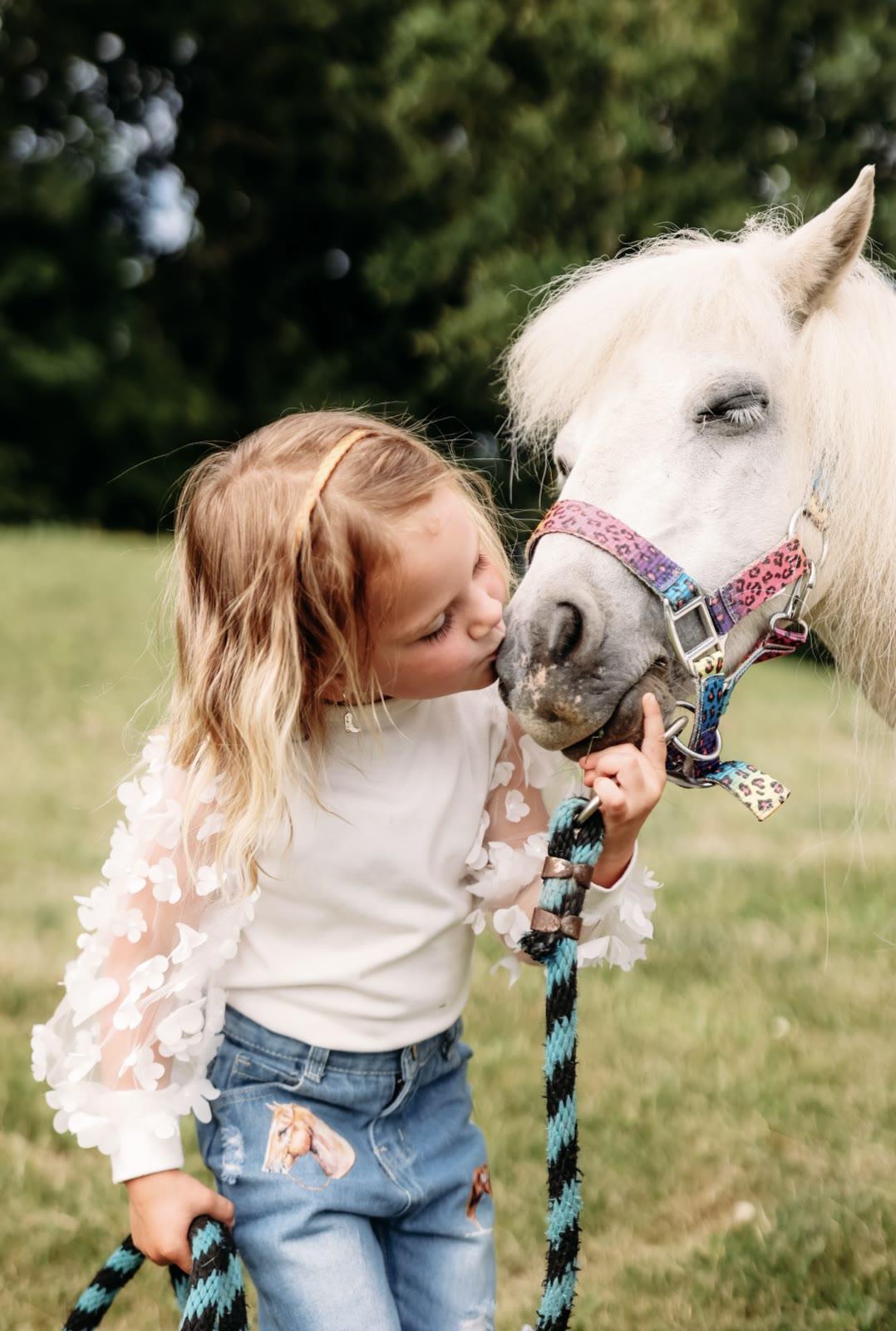 A young girl with a headband kisses the nose of a white pony wearing a colorful halter, holding its lead rope in a grassy field.
