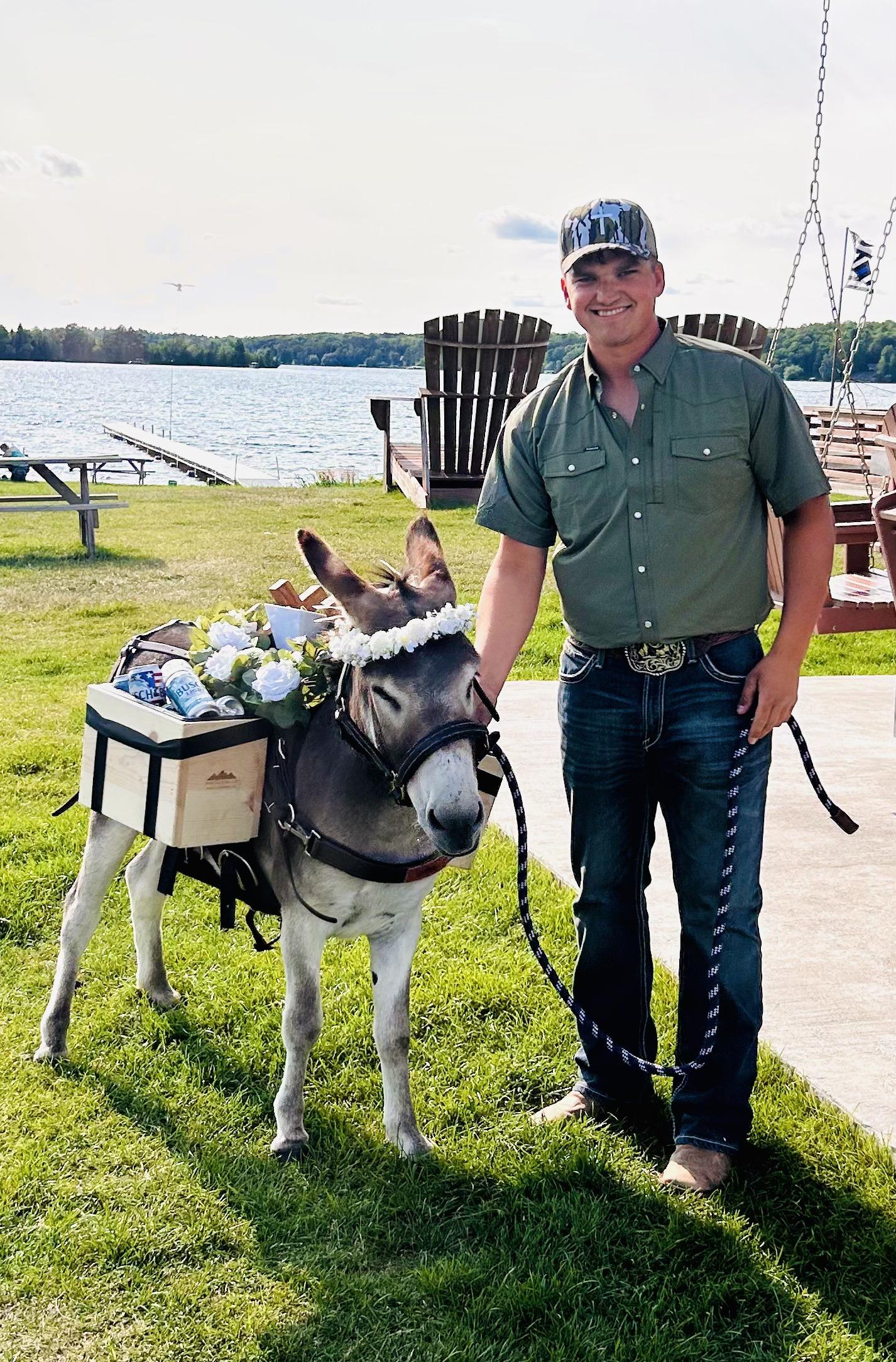 Kyle Knops standing lakeside with Guapo, a grey miniature donkey wearing a white flower crown and a wooden saddle bar stocked with cold cans.