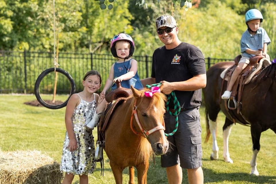 Kyle Knops steadying a young rider on a saddled pony at a backyard party