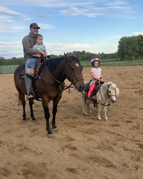 Duke, a bay quarter horse, with Kyle Knops in the saddle holding a small child, next to a white pony in the farm arena.